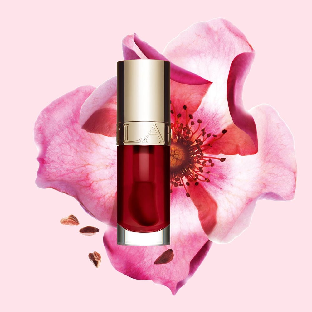 Packshot of Clarins Lip Comfort Oil in Cherry, placed next to a sweetbriar rose on a light pink background.