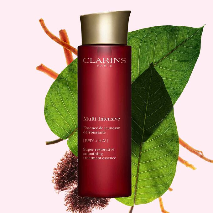 Packshot of Clarins Super Restorative Treatment Essence near red jania, Harungana leaves, and ginseng root on pink backdrop.