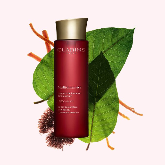 Packshot of Clarins Super Restorative Treatment Essence near red jania, Harungana leaves, and ginseng root on pink backdrop.